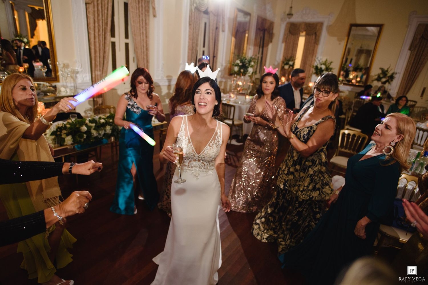 Bride celebrates on the dance floor surrounded by guests holding colorful glow sticks at wedding.