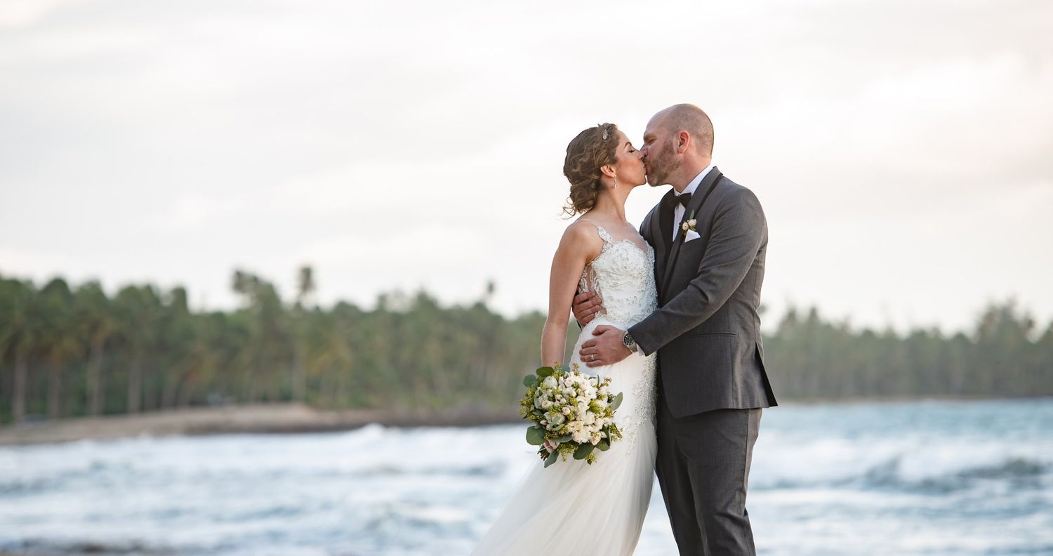 Bride and groom kissing on a tropical beach with ocean waves and palm trees in the background.