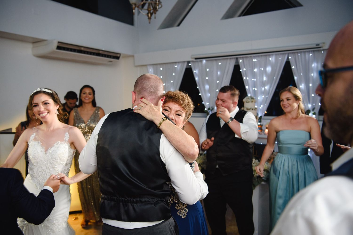 Wedding reception dance floor with bride in white gown, guests dancing and celebrating under twinkling fairy lights.