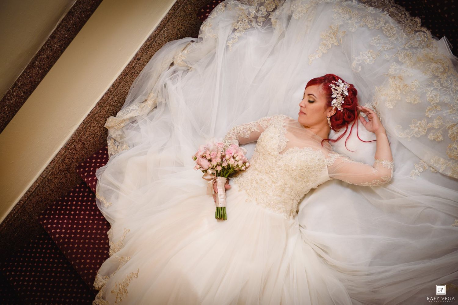 Bride in elegant lace ball gown with red hair and floral accessories, holding pink bouquet while lying on stairs.