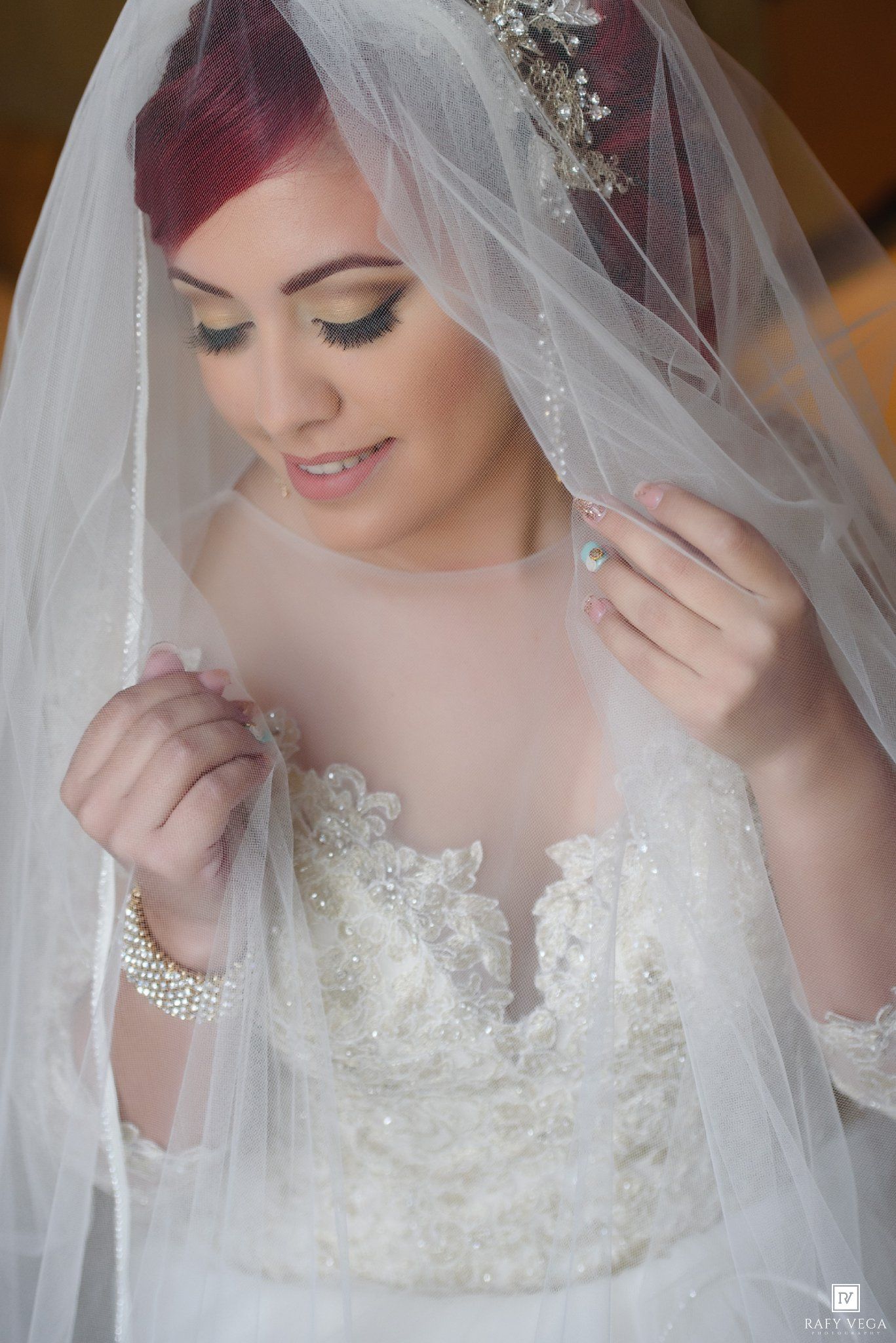 A bride with red hair wearing a lace wedding gown and crystal tiara, delicately holding her sheer white veil.