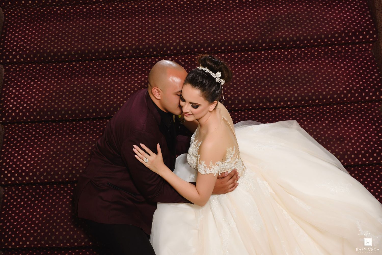 Bride in white off-shoulder gown and groom in burgundy suit share a tender embrace on red staircase.