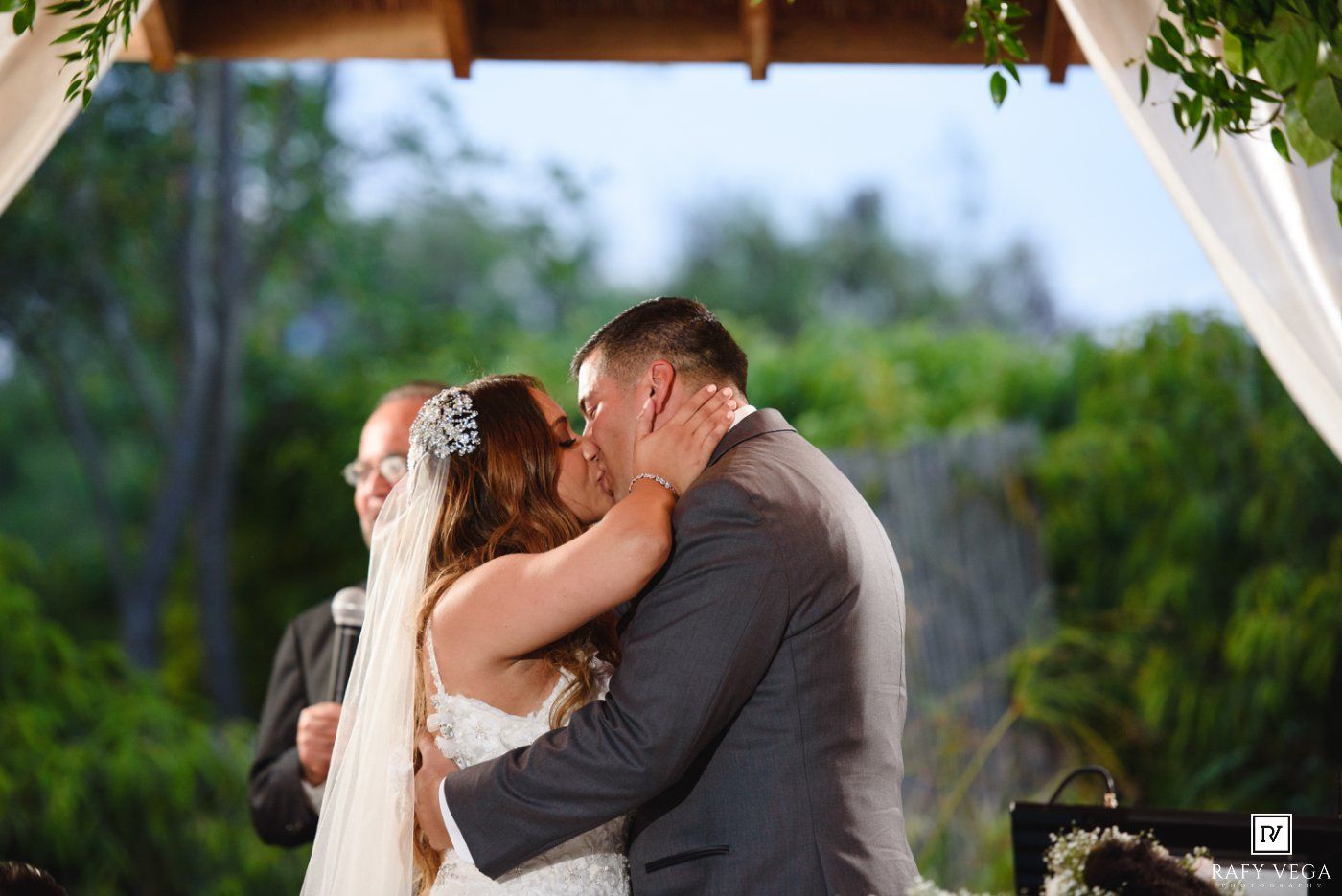 Bride and groom share first kiss at outdoor wedding ceremony under floral arch with lush green tropical backdrop.