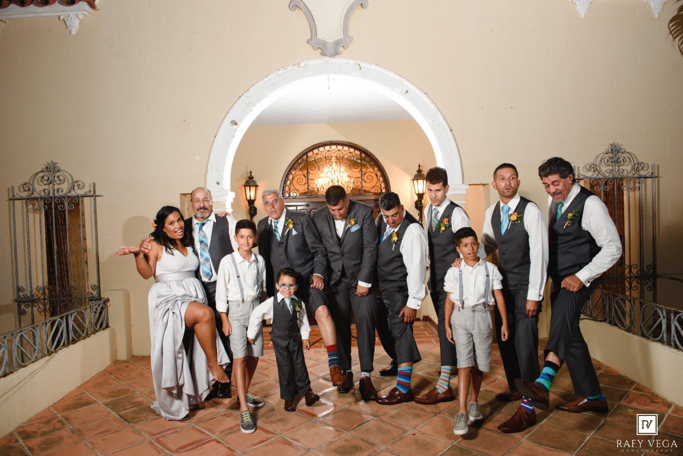 Wedding party poses under arched doorway, groomsmen in gray vests with bride in white dress at elegant venue.