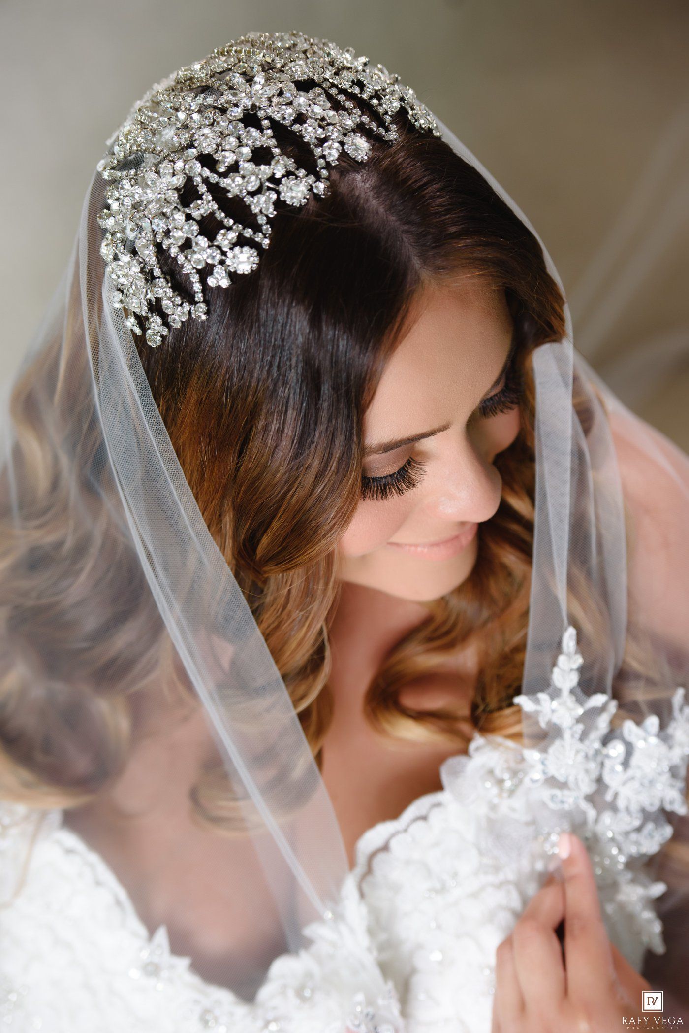 Bride wearing an ornate crystal tiara with a flowing white veil and lace wedding dress, looking down gracefully.