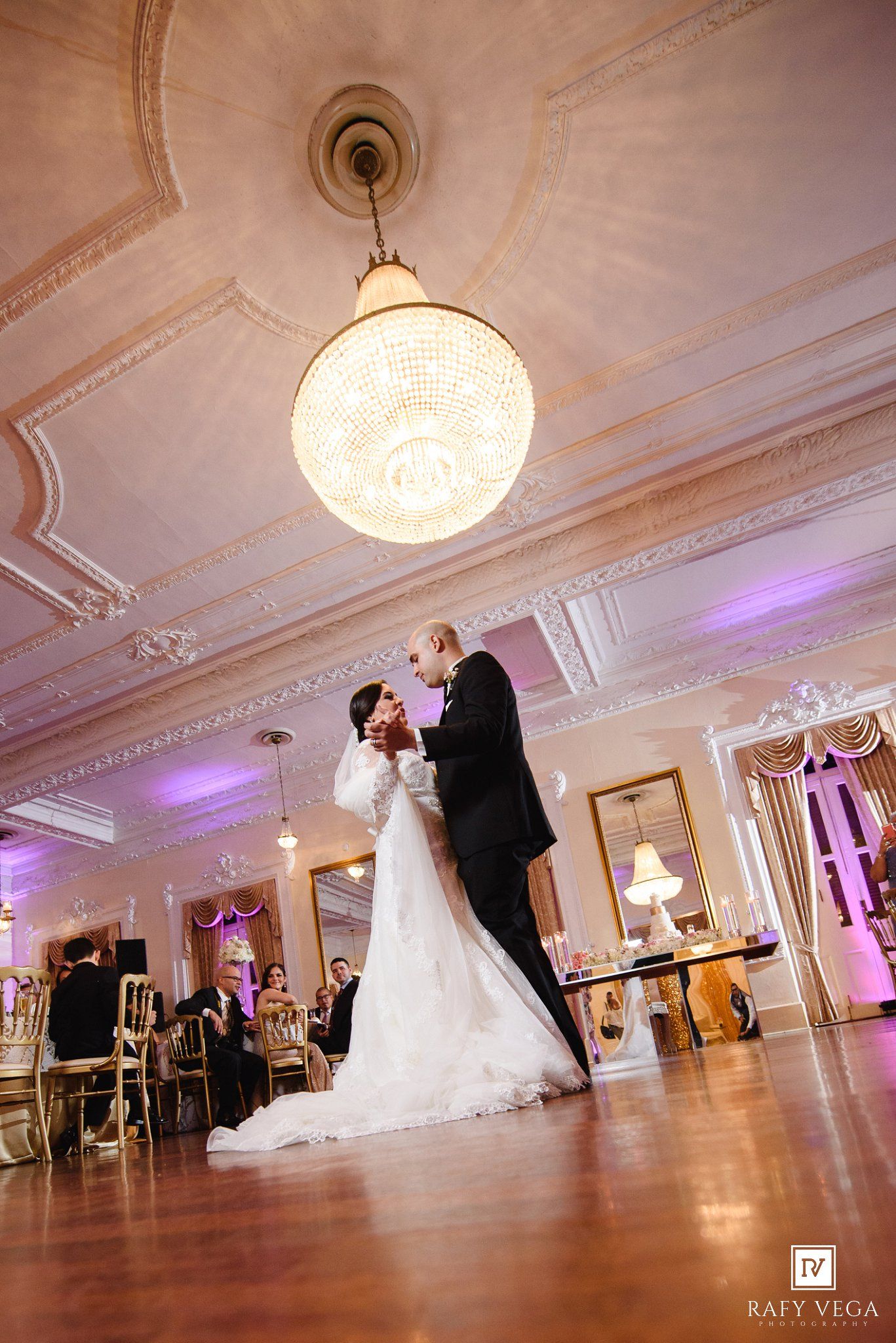 Bride and groom share their first dance under a crystal chandelier in an elegant ballroom with purple uplighting.