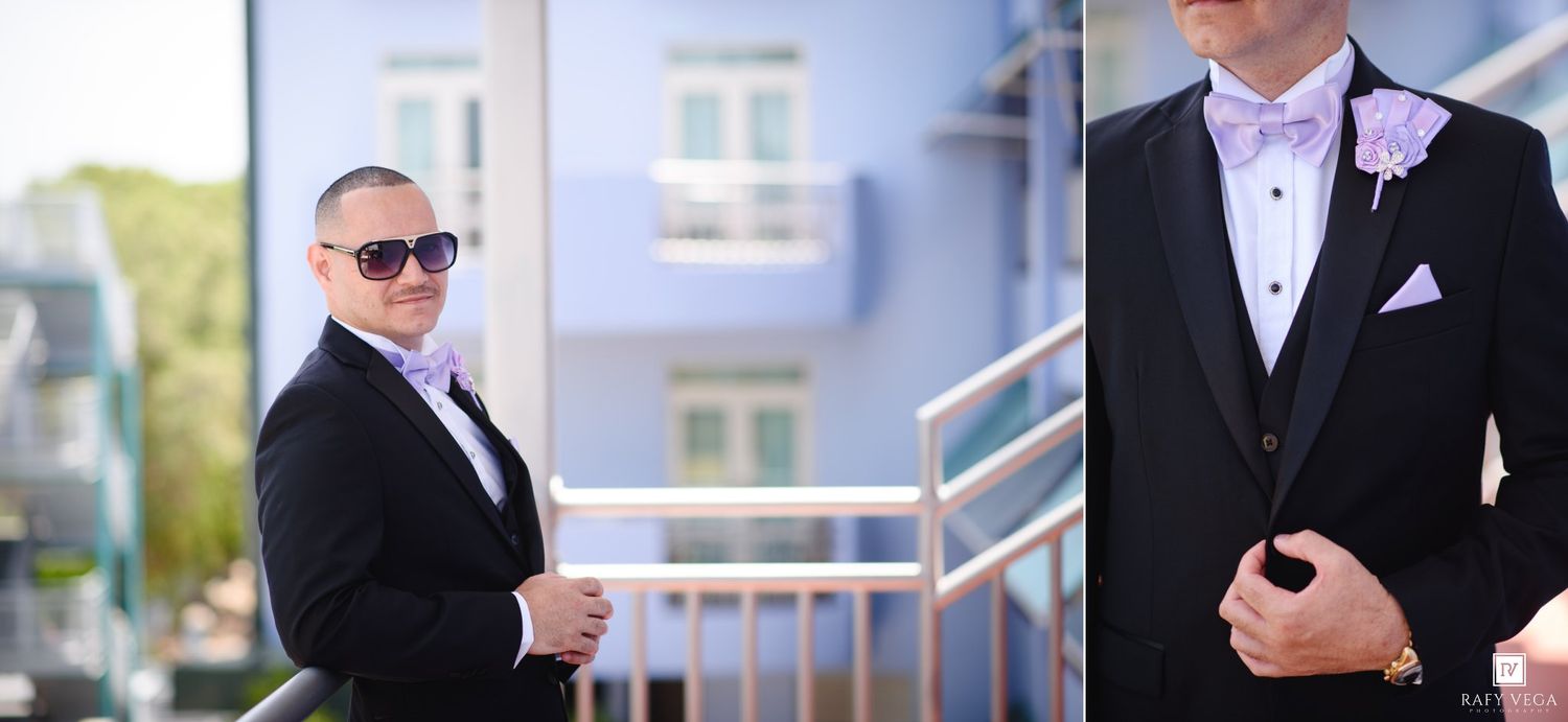Groom in black tuxedo with lavender bow tie and boutonniere poses outdoors near building steps on wedding day.