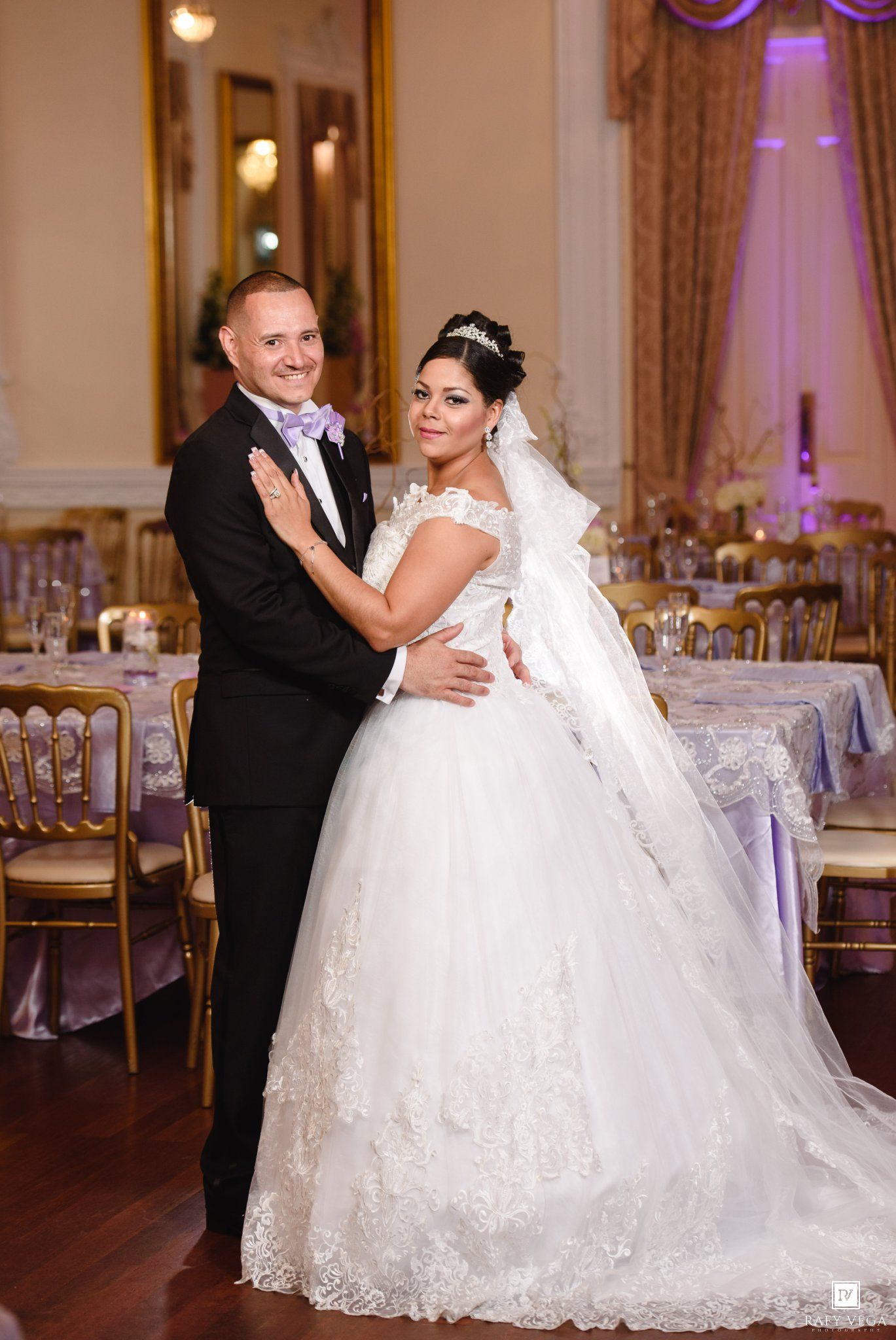 Bride in white ballgown and groom in black tuxedo embracing at elegant wedding reception venue.