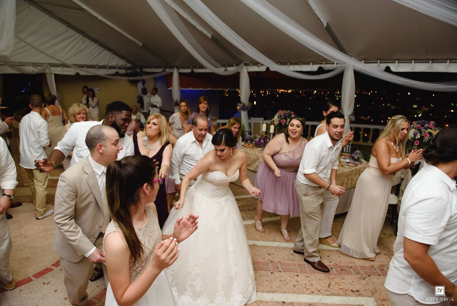 Bride and wedding guests dance joyfully under a white tent at an outdoor evening wedding reception.