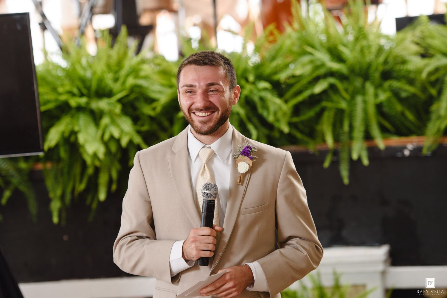 Smiling groom in tan suit with purple boutonniere holds microphone while giving wedding speech near lush green ferns.