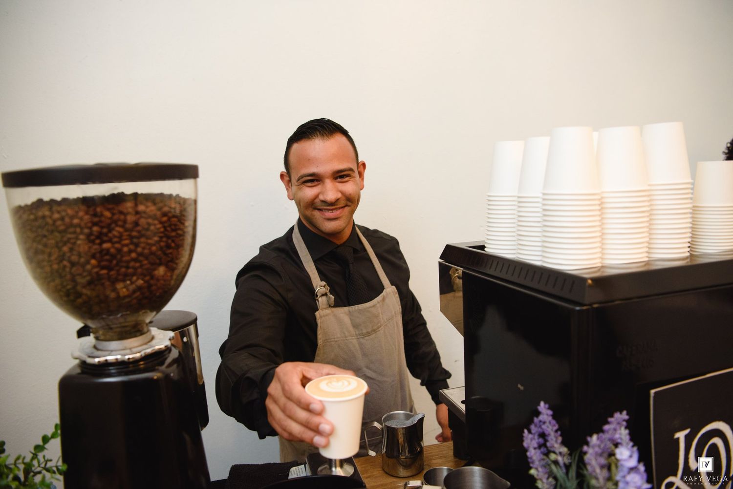 Smiling barista in apron serving a latte at a coffee bar with bean grinder and stacked cups nearby.
