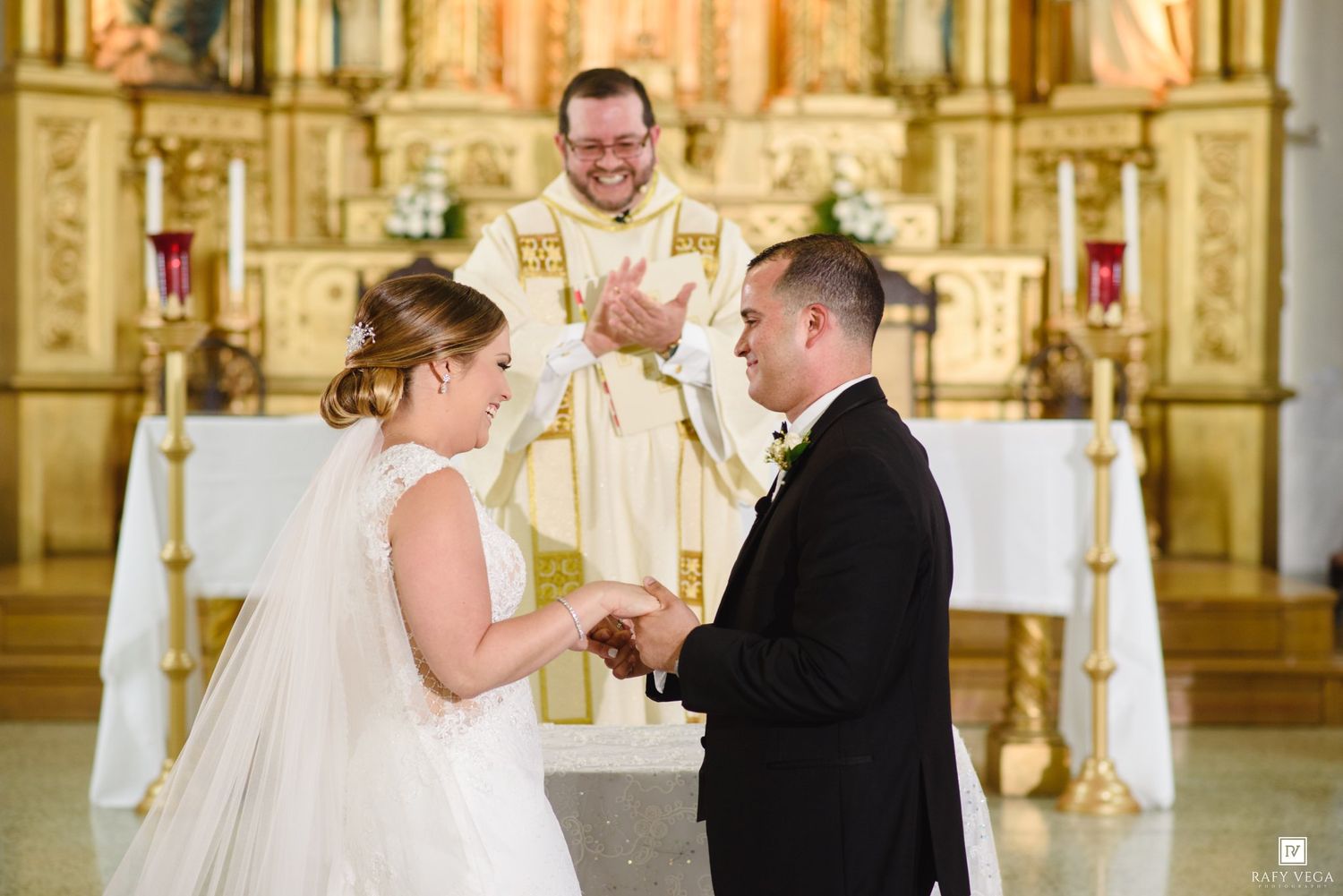 Priest officiating wedding ceremony as bride and groom exchange rings at ornate golden altar.