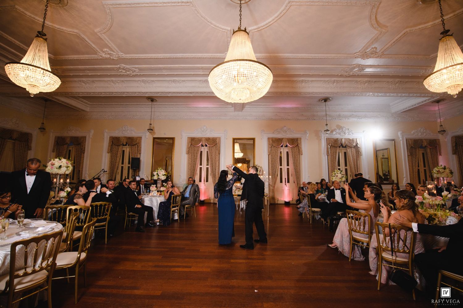 Elegant wedding reception in a grand ballroom with chandeliers, gold chairs, and couple sharing first dance.