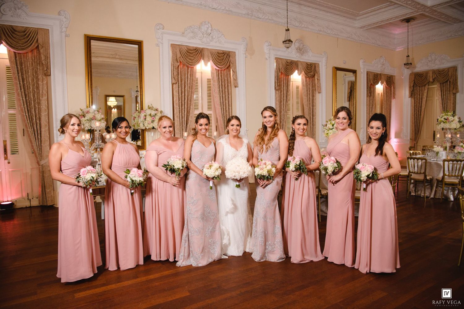 Bride poses with eight bridesmaids in matching dusty rose gowns holding bouquets in a grand ballroom.