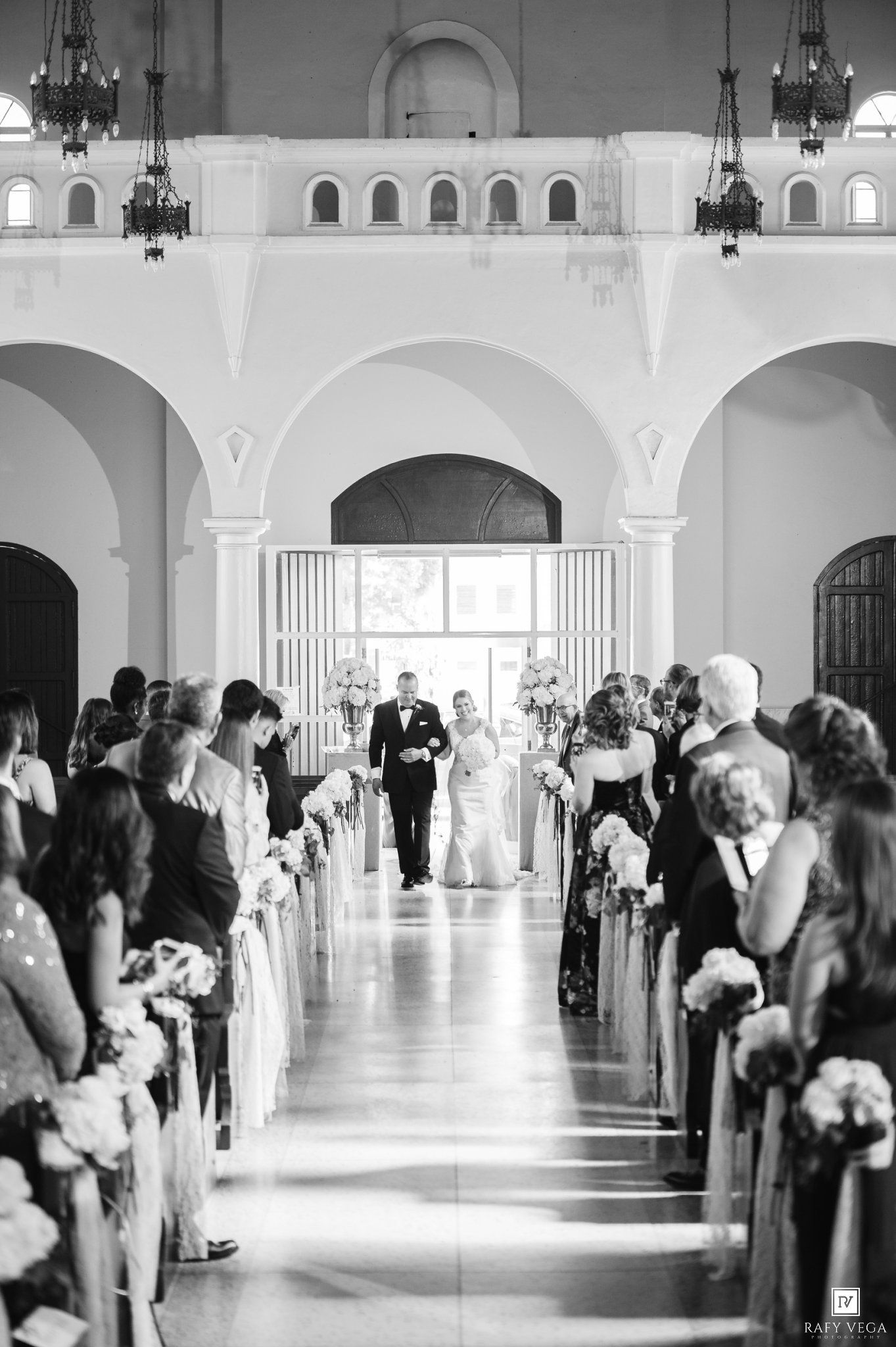 Black and white wedding ceremony photo of bride walking down aisle in elegant church with arched ceilings and floral decorations.