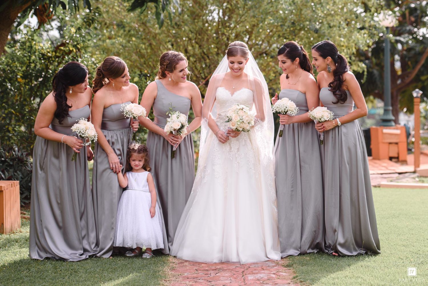 Bride in white gown with bridesmaids in gray dresses and flower girl in white, holding bouquets outdoors.