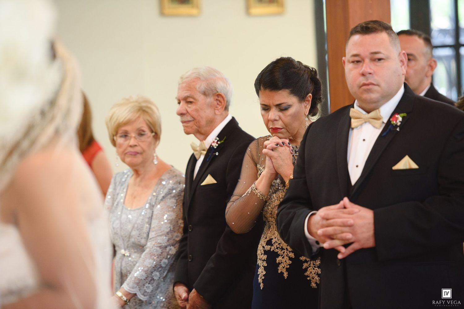 Wedding guests praying during ceremony, elegant attire including tuxedo and formal gowns.
