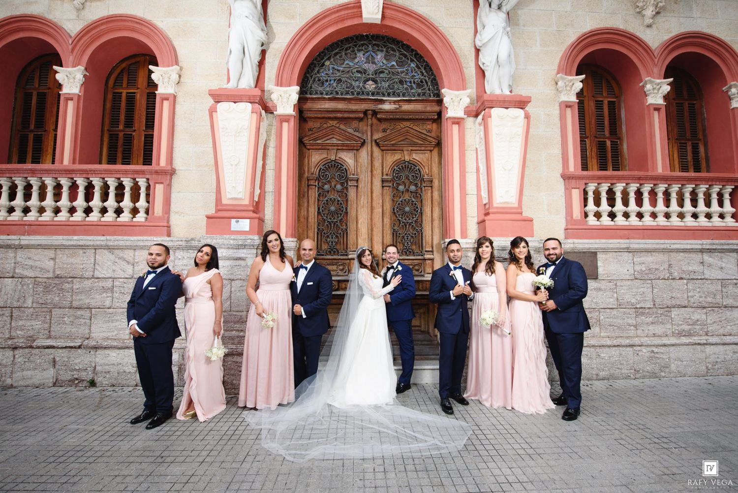 Wedding party poses in blush gowns and navy suits before ornate pink building with wooden doors.