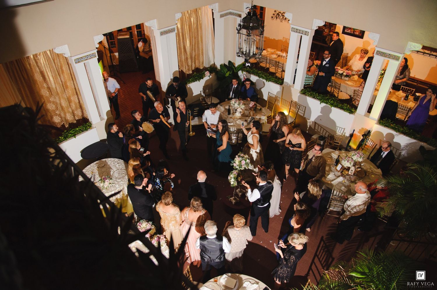 Aerial view of elegant wedding reception with guests mingling on dance floor in decorated venue with columns.