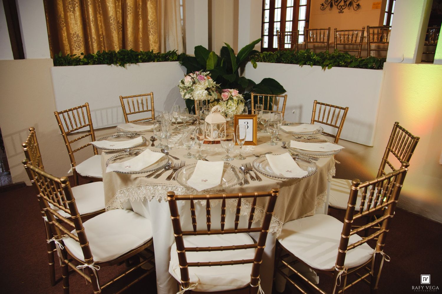 Elegant round banquet table with gold Chiavari chairs, ivory linens, and floral centerpiece in a decorated event venue.