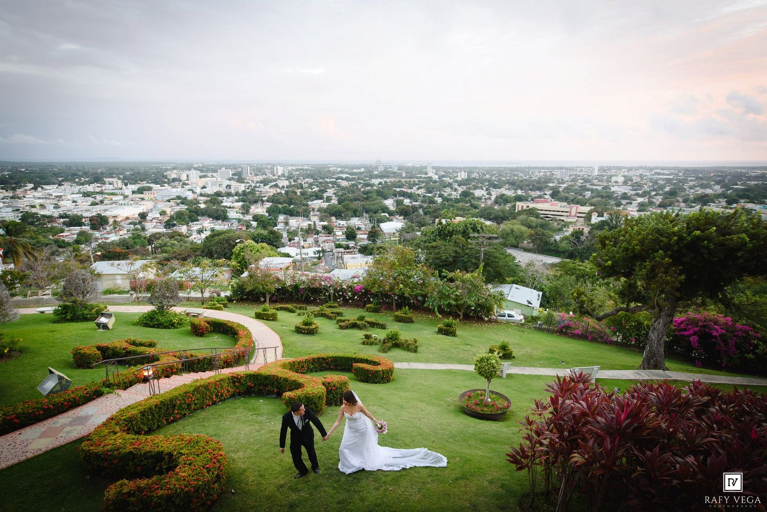 Bride and groom walking through lush garden venue with panoramic city views at sunset wedding celebration.