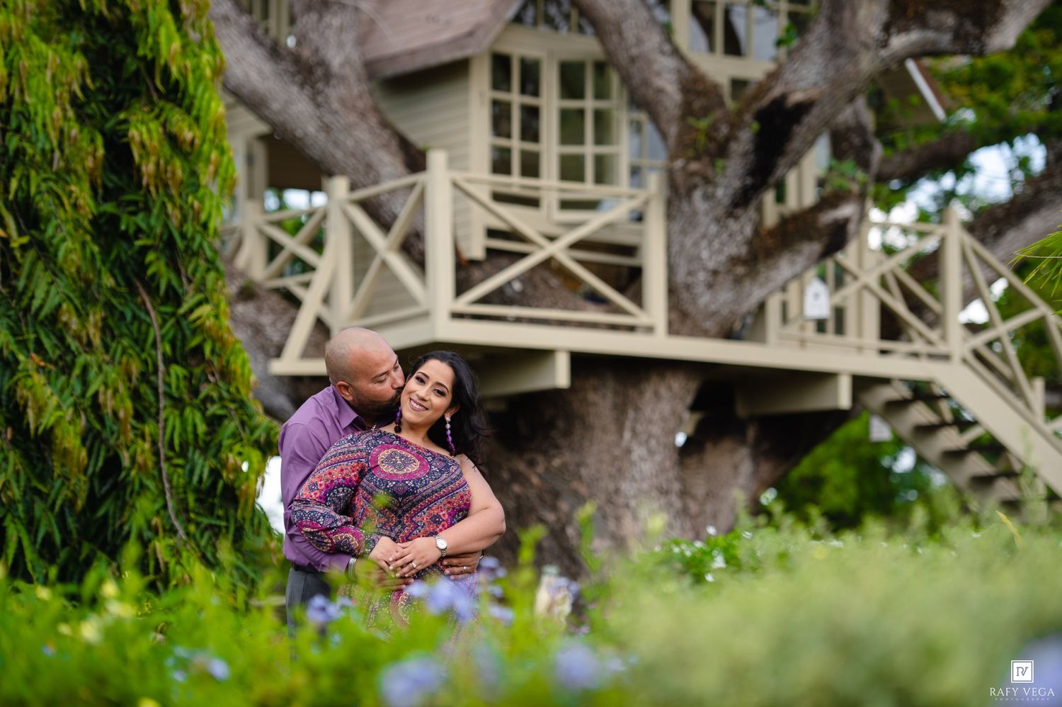 Couple embracing romantically in a lush garden with a charming treehouse in the background.