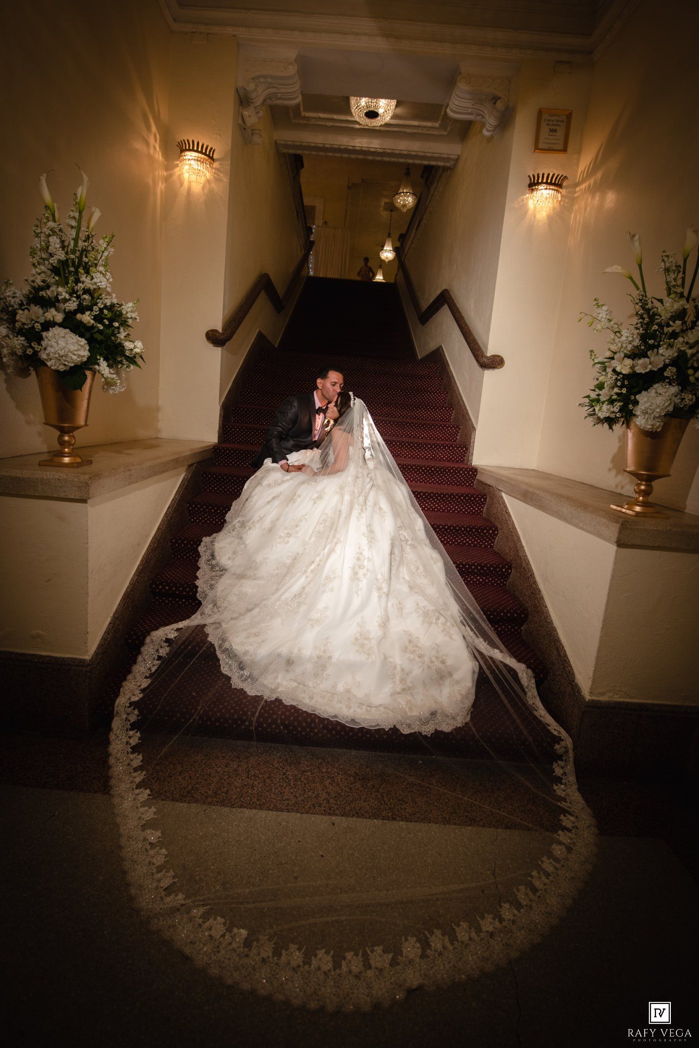 Bride and groom share a romantic moment on a grand staircase, her voluminous gown and lace veil cascading down the steps.