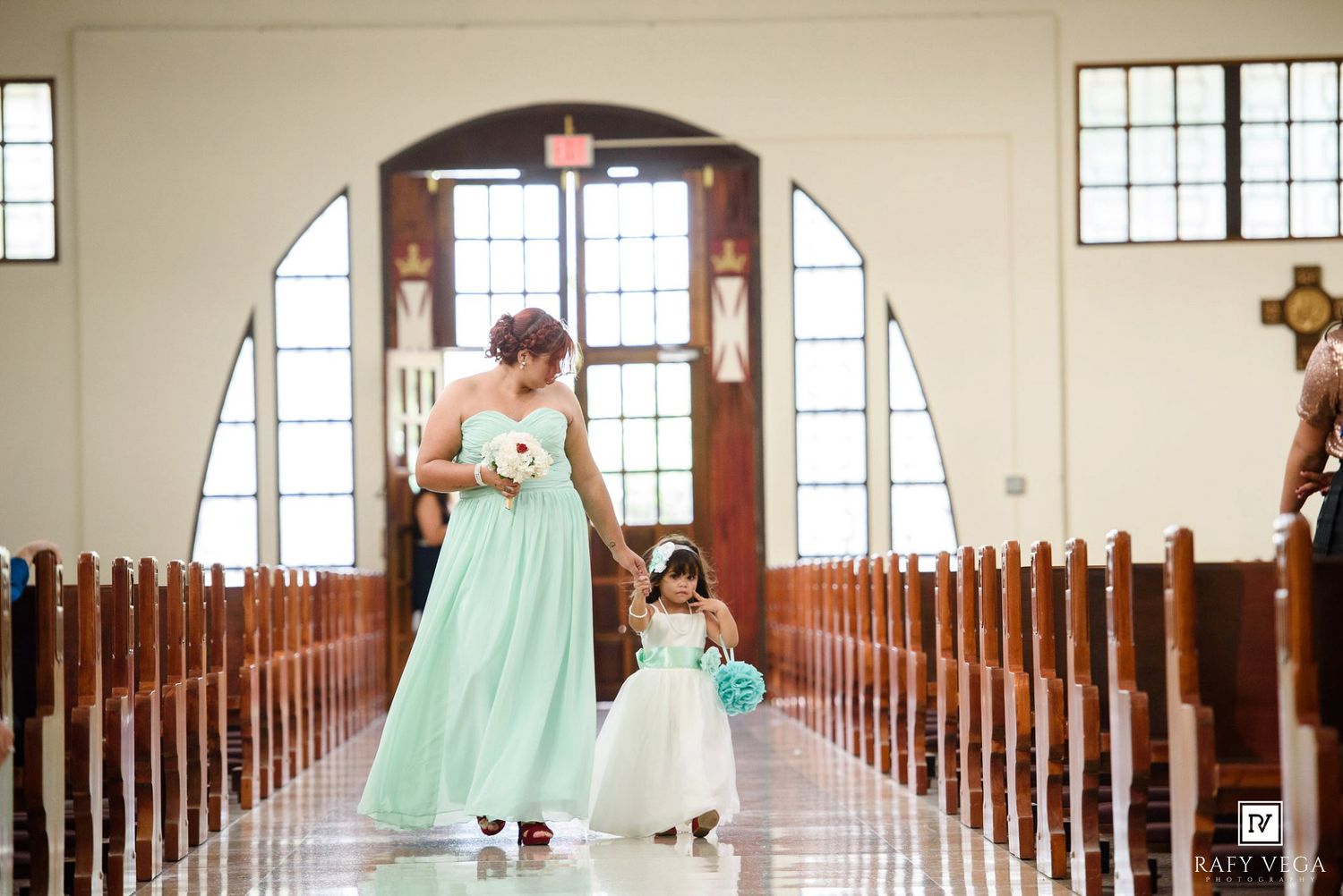Bridesmaid in mint green gown walks with flower girl down church aisle during elegant wedding ceremony.