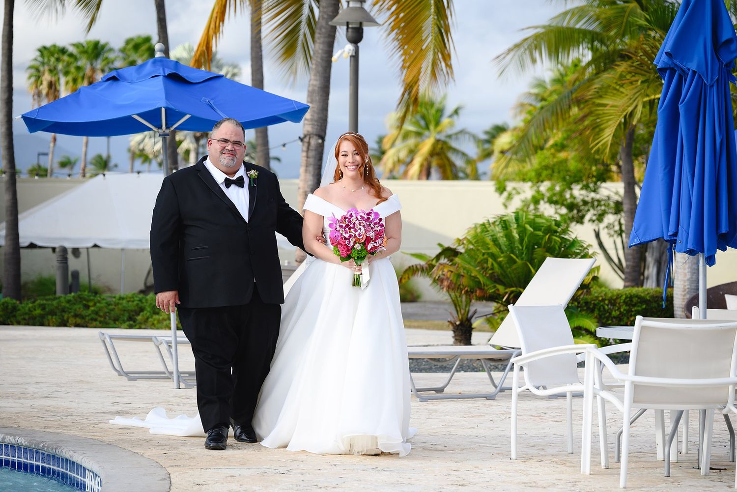 Bride and groom posing poolside at a tropical resort wedding, with palm trees and blue umbrellas in the background.