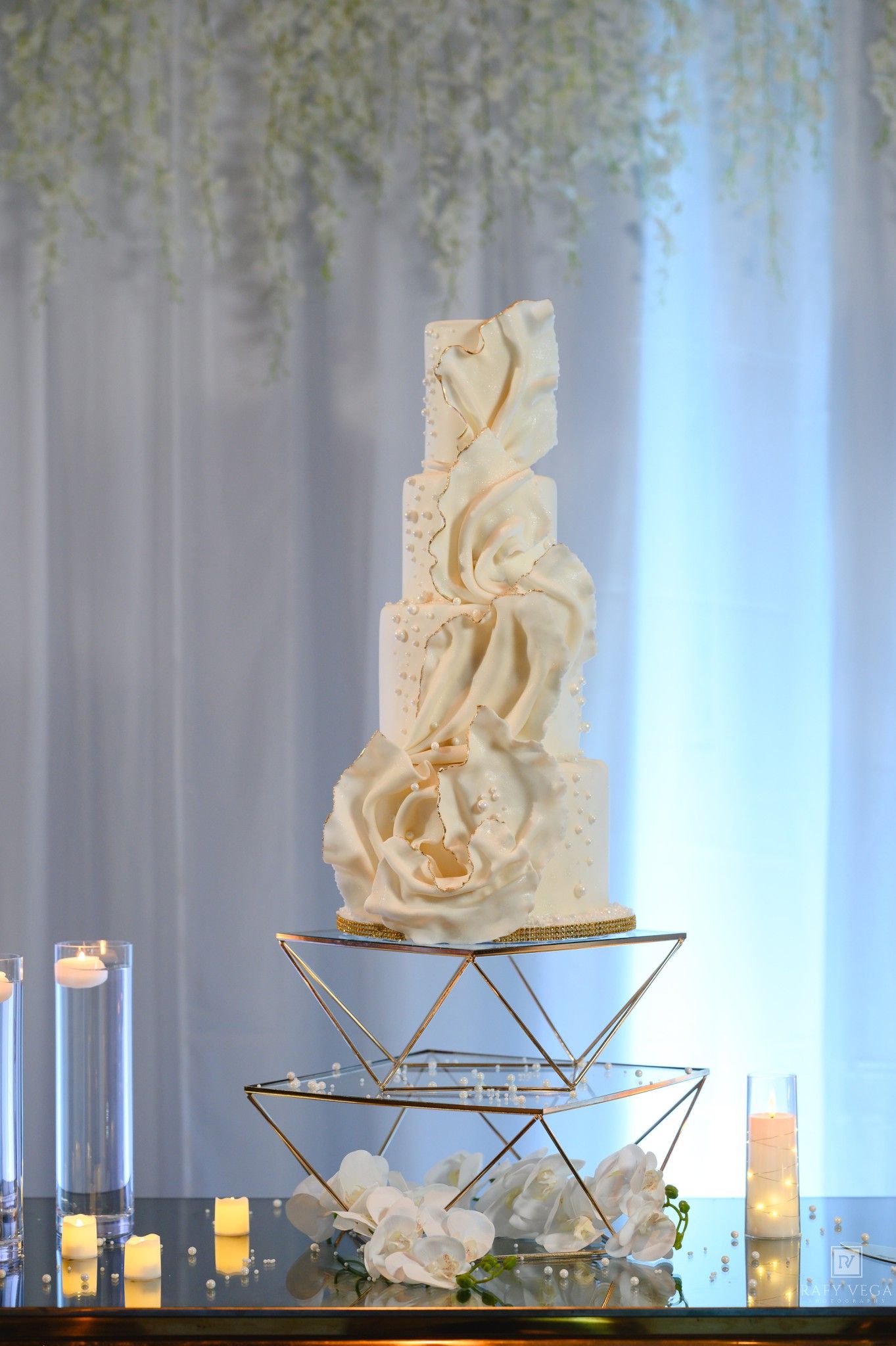 Elegant multi-tier white wedding cake with ruffles and gold accents on geometric stand, flanked by candles and white flowers.