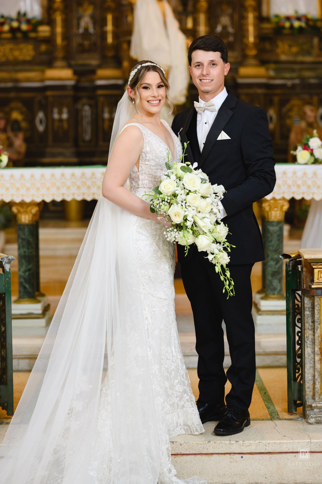 Bride in white lace gown holding cascading bouquet stands with groom in black tuxedo at church altar.