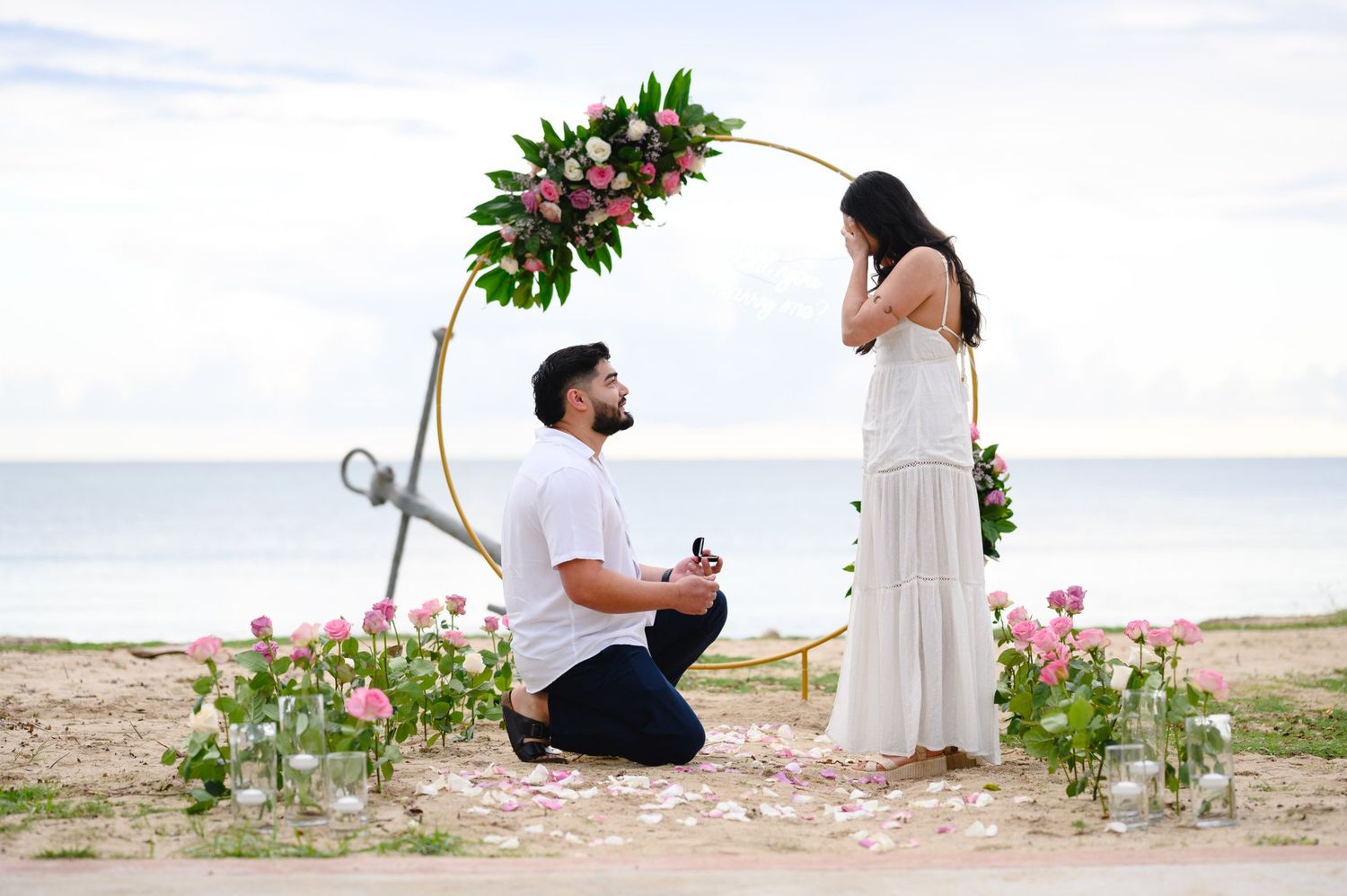 Man proposes to woman on beach with floral arch, rose petals, and ocean backdrop.