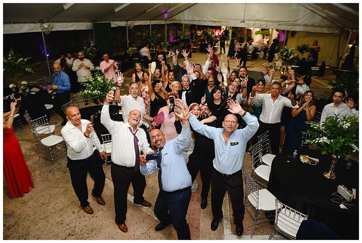 Wedding guests celebrate with raised glasses and cheers at a festive outdoor reception under a white tent.
