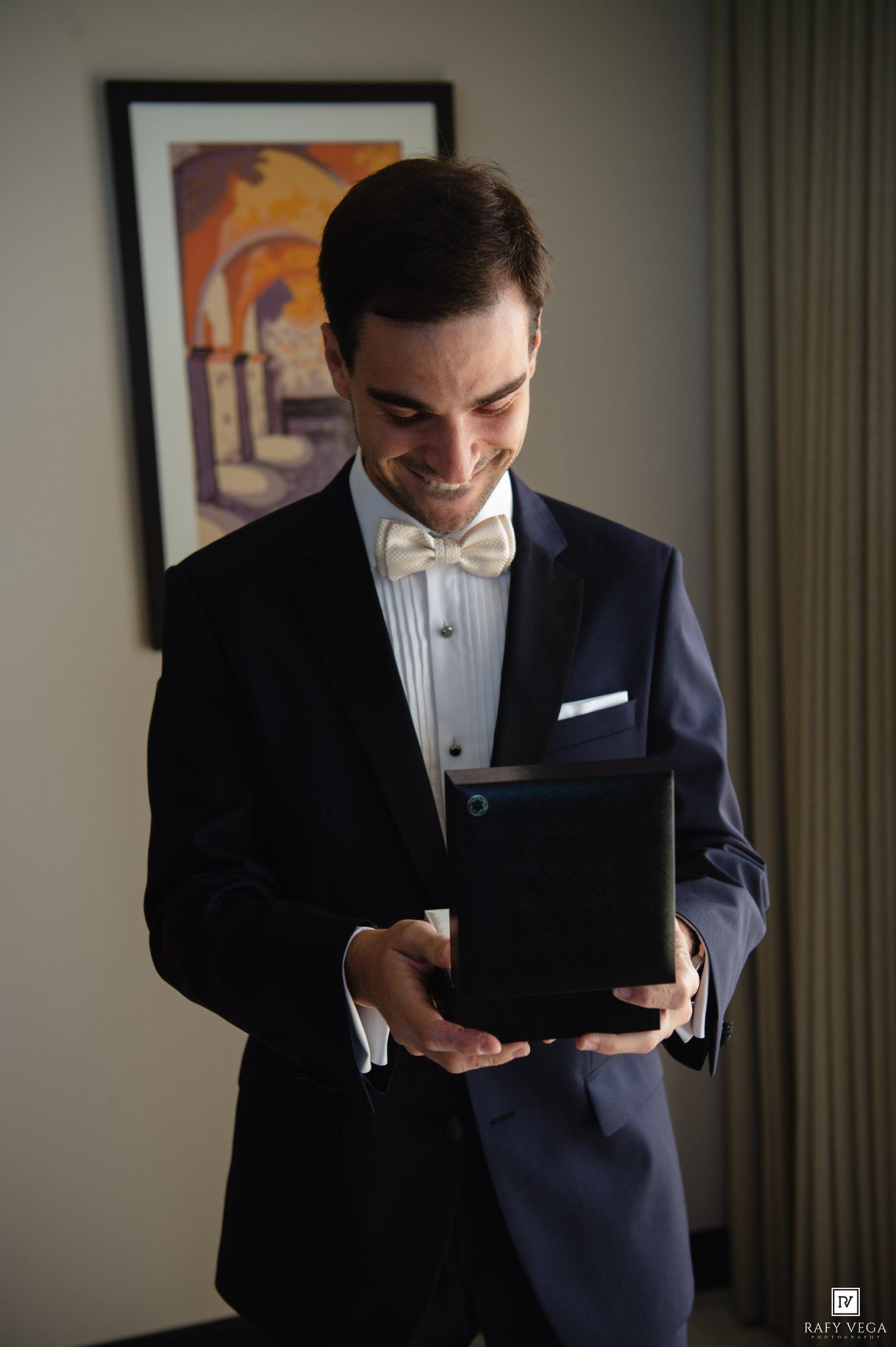 Smiling groom in navy tuxedo with white bow tie opening a gift box on his wedding day.