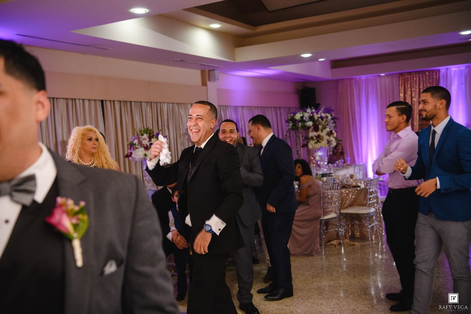 Guests celebrating at an elegant wedding reception hall with purple lighting and floral decorations.