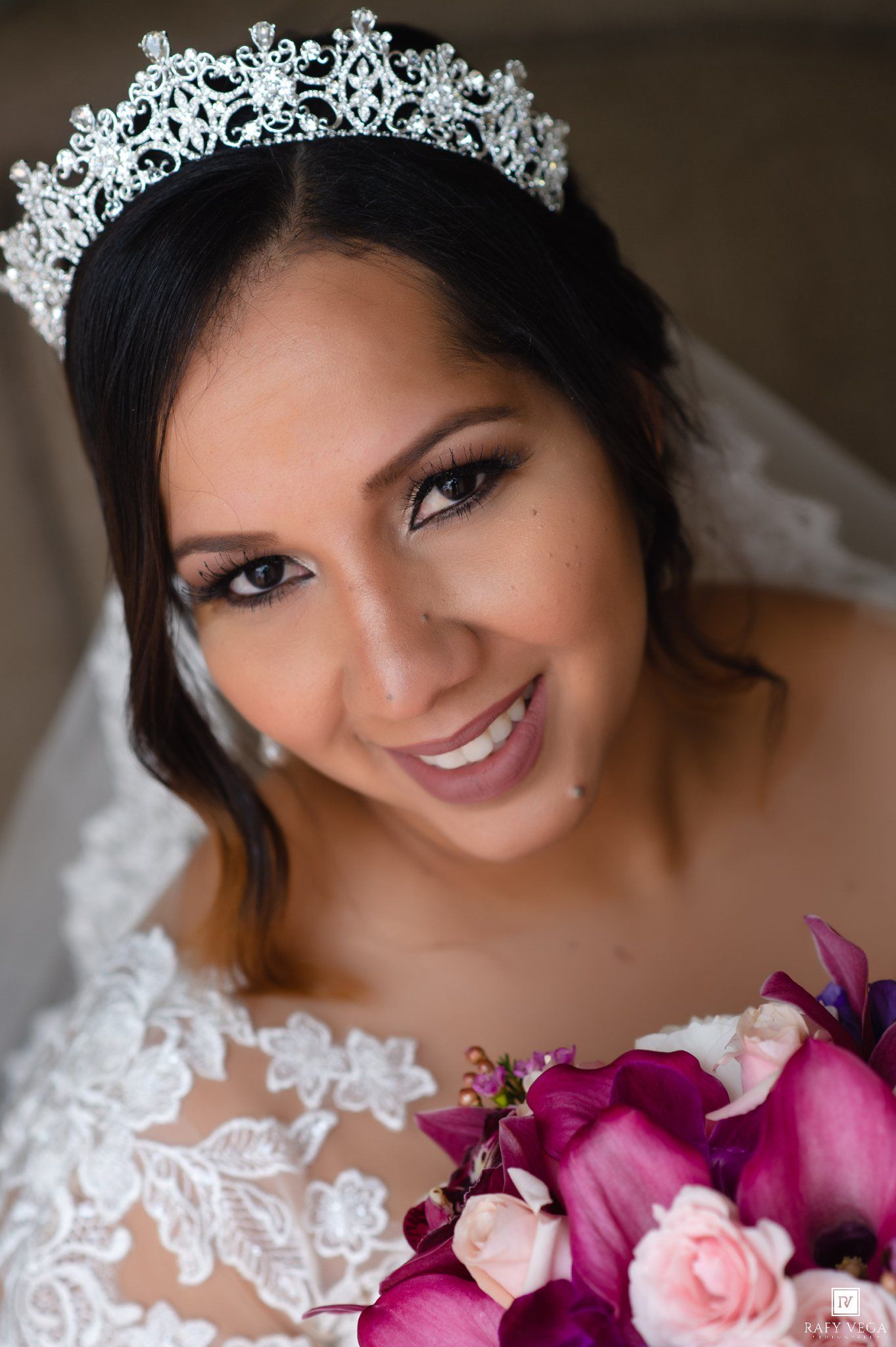 Smiling bride wearing a crystal tiara and lace wedding dress holds a bouquet of purple orchids and pink roses.