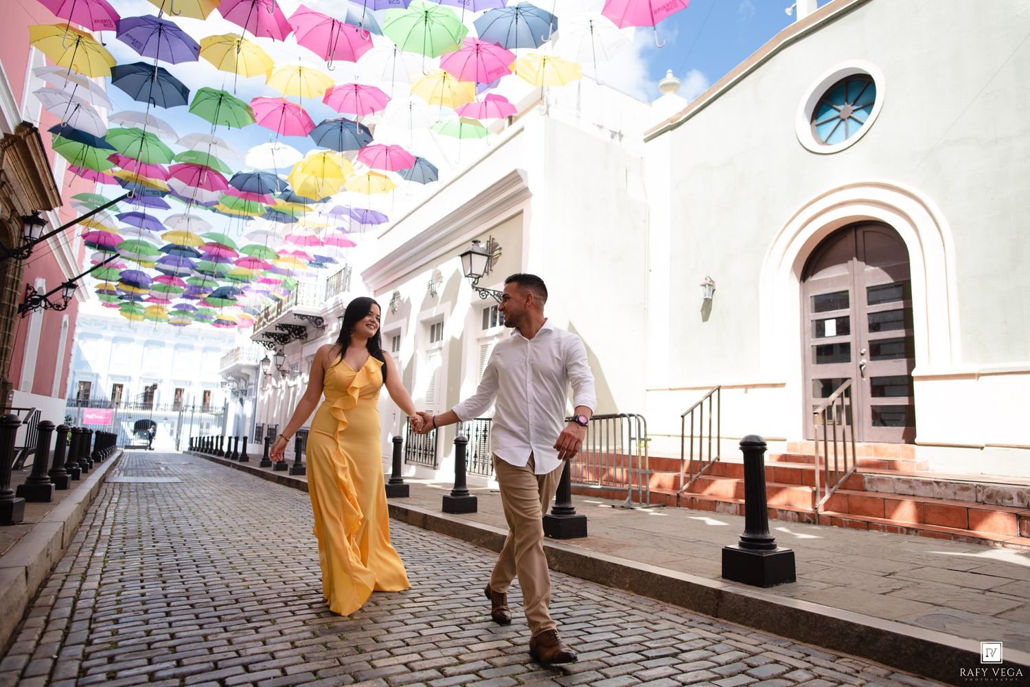 Couple holding hands walking under colorful umbrella canopy on cobblestone street in Old San Juan, Puerto Rico.