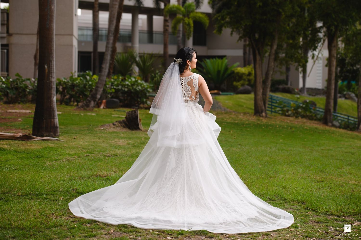 Bride in elegant white layered wedding gown with lace back and long veil standing in lush green garden.