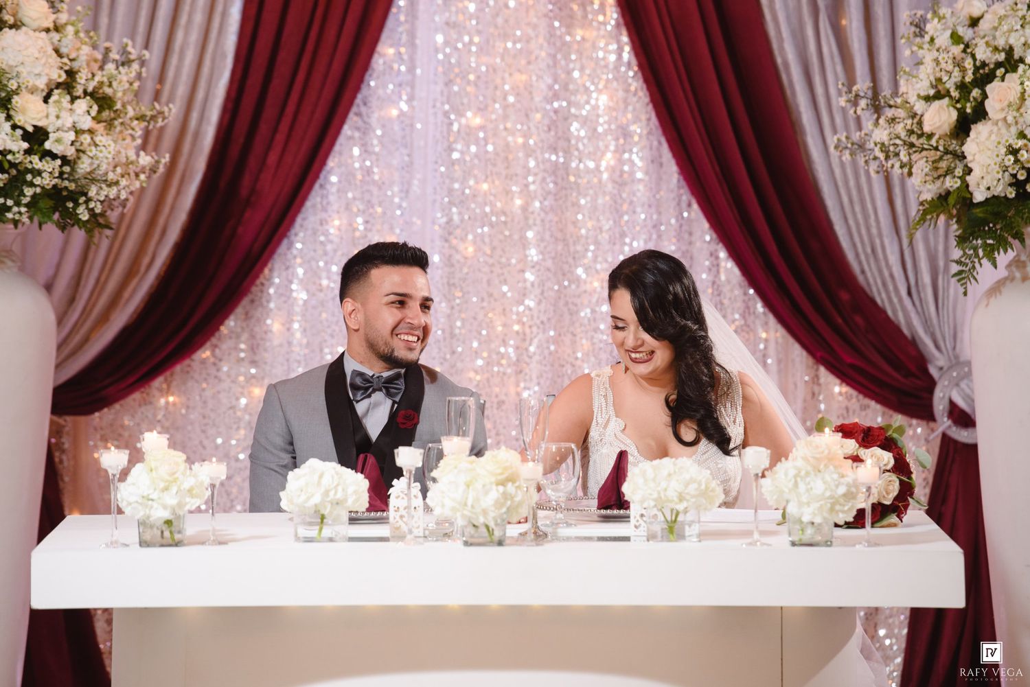 Bride and groom smiling at sweetheart table decorated with white flowers and candles at elegant wedding reception.