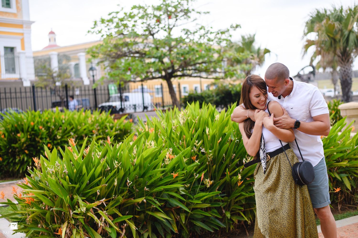 Couple embracing in a lush tropical garden plaza with palm trees and colorful buildings in the background.
