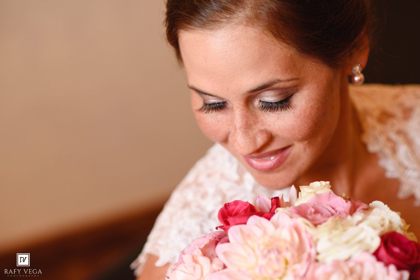 Bride smiling softly at her pink and white wedding bouquet, wearing a lace dress with elegant makeup.