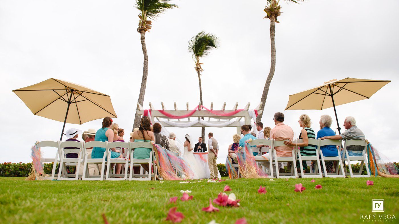 Guests seated at an elegant outdoor tropical wedding ceremony with palm trees and pink decorations under cloudy skies.