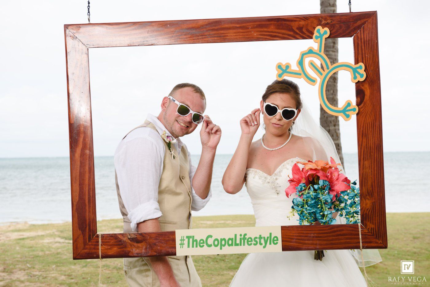 Bride and groom posing in a decorative photo frame on a beach with #TheCopaLifestyle hashtag at their wedding.