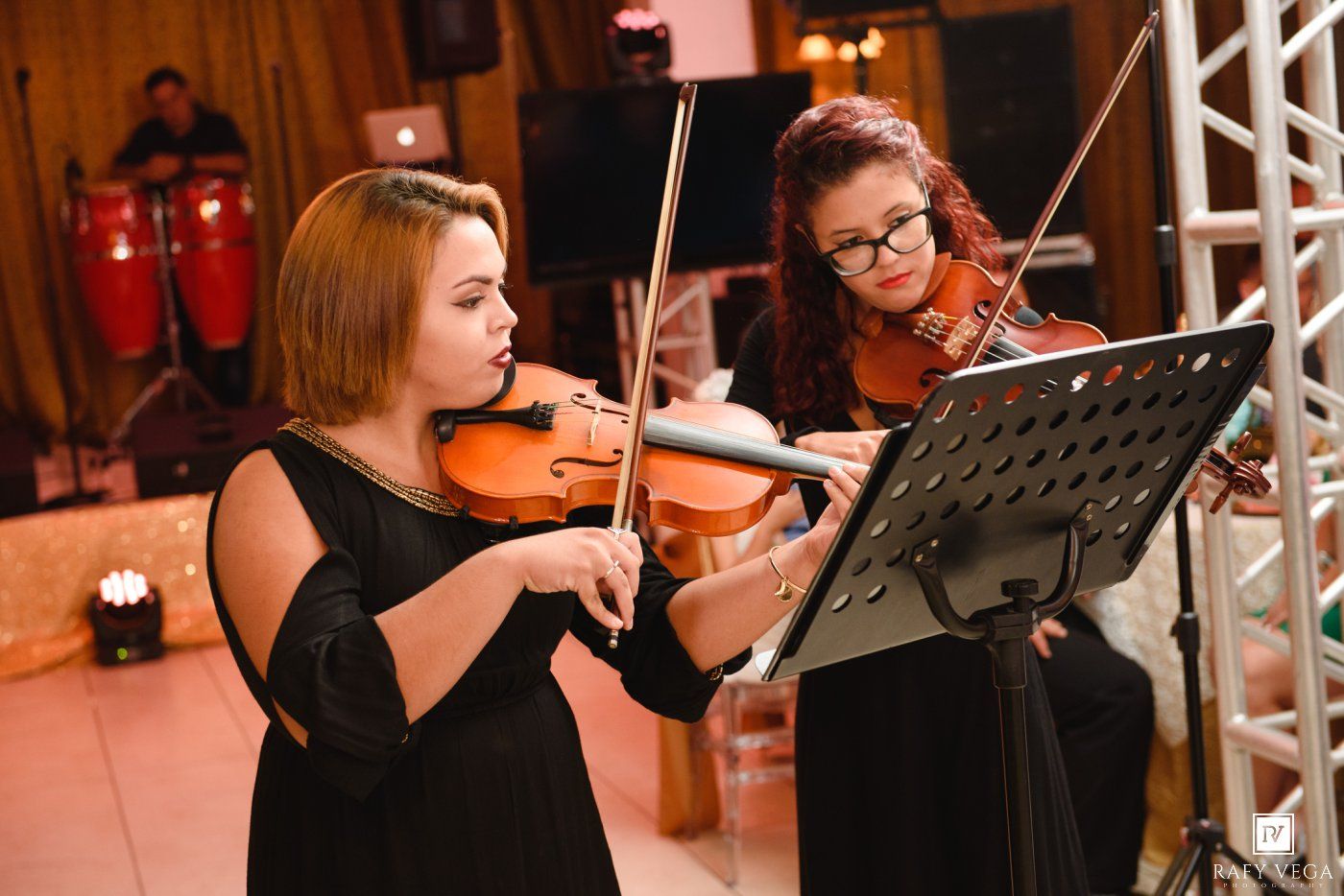 Two women in black dresses playing violins at a formal event with sheet music stands.