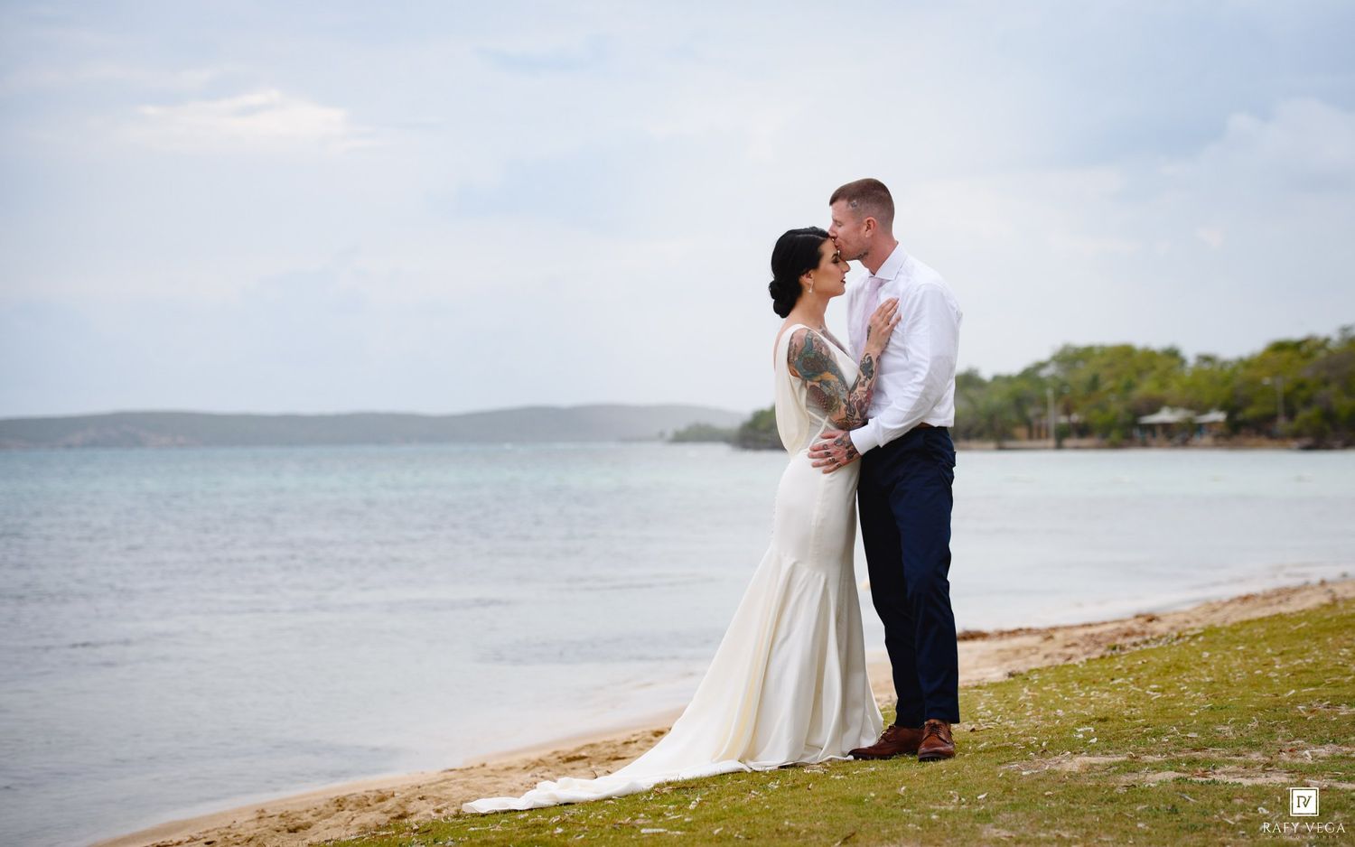 Bride and groom share a romantic moment on a sandy beach shoreline with calm ocean waters and trees in the background.