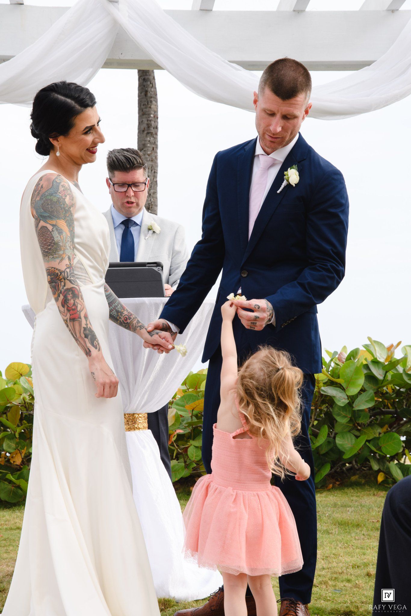 Tattooed bride and groom hold hands at outdoor wedding ceremony while flower girl in pink dress reaches up.