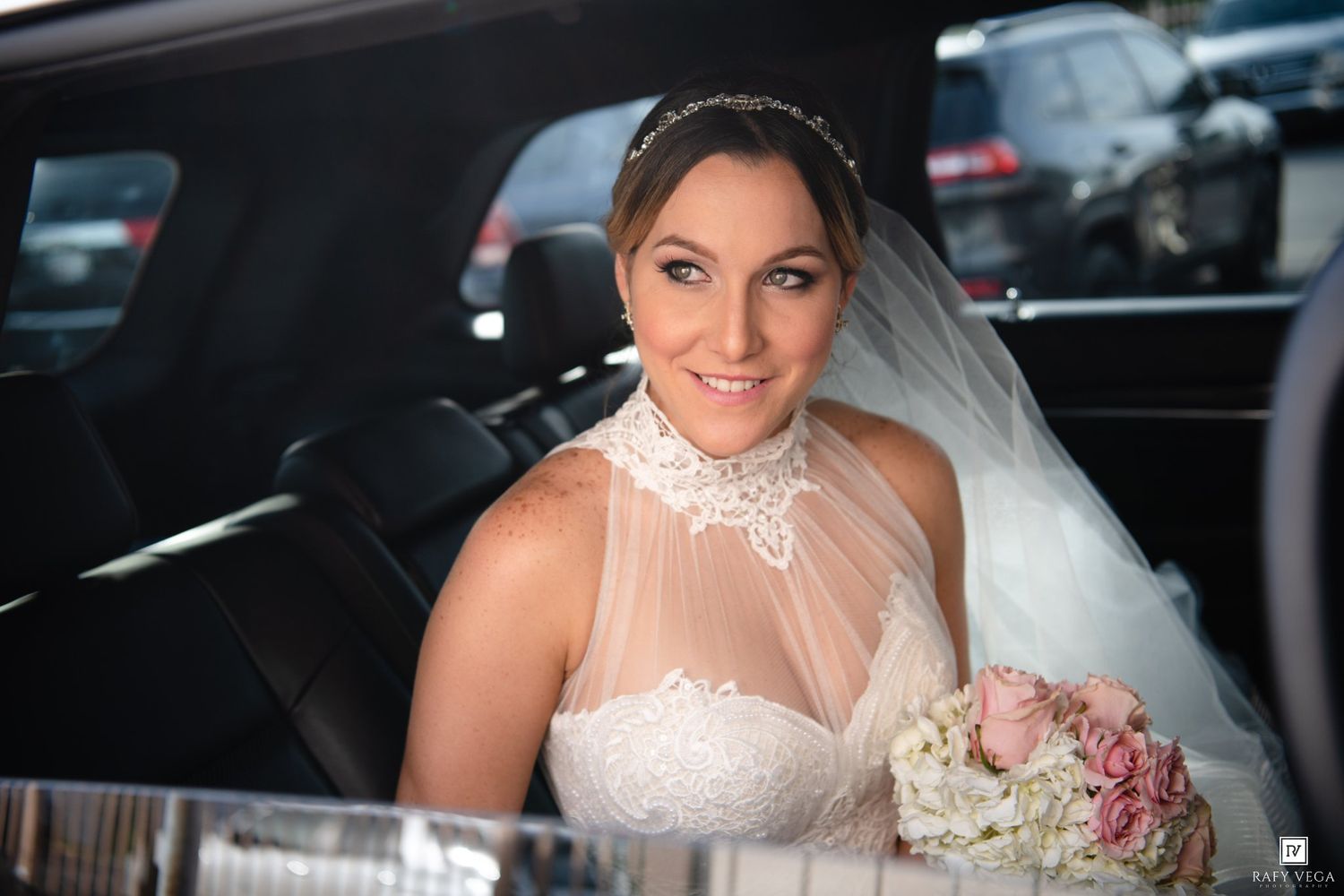 Smiling bride in white lace wedding dress and veil holding bouquet while seated in a luxury limousine.