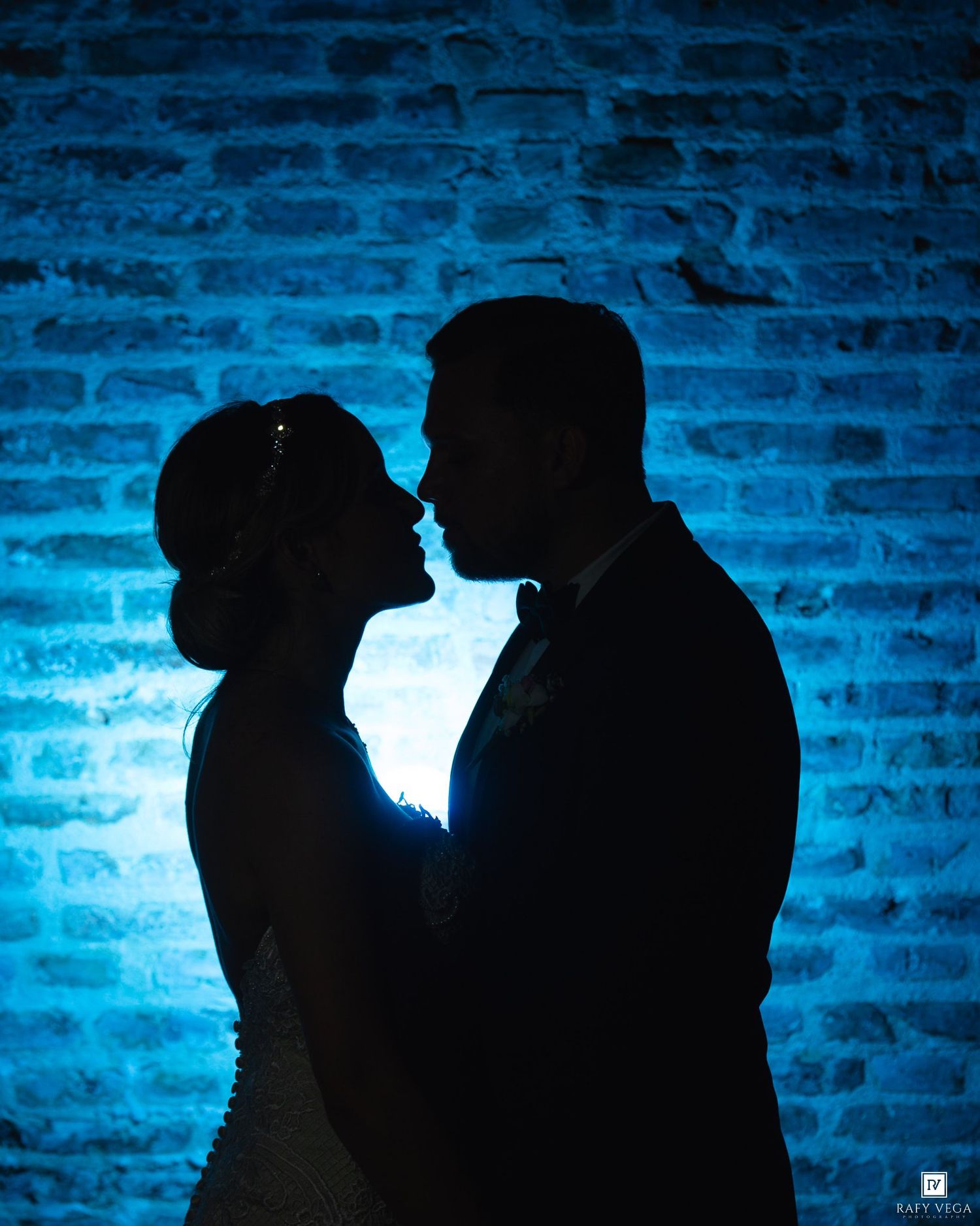 Romantic silhouette of bride and groom almost kissing against a blue-lit brick wall.