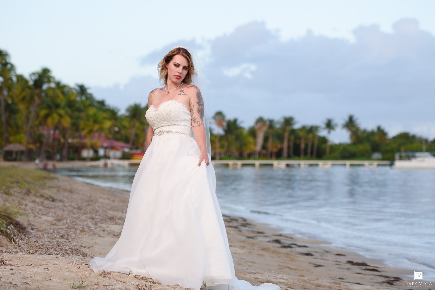 Bride in a strapless white wedding gown posing on a tropical beach with palm trees and calm water at dusk.