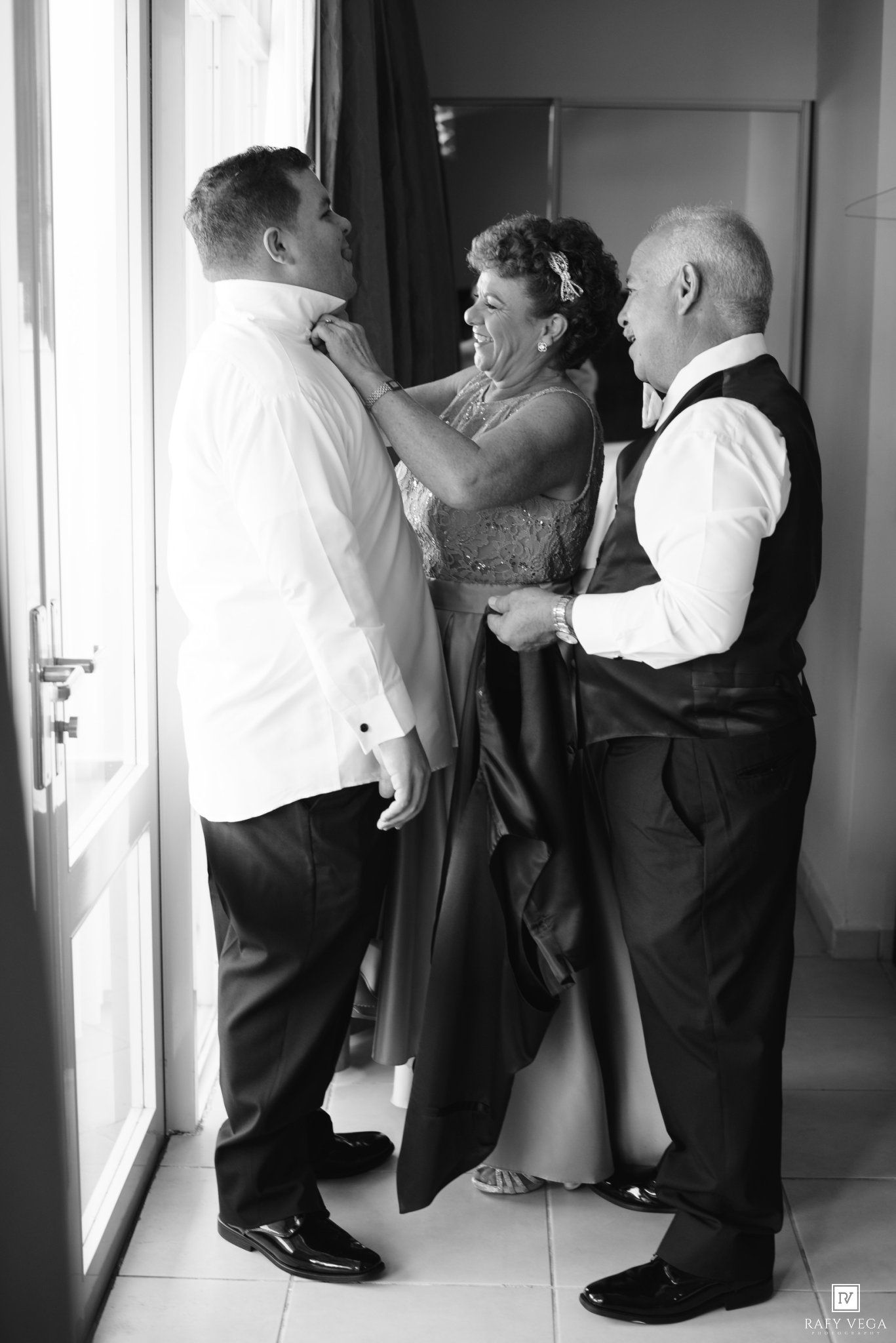 Black and white wedding photo of groom getting tie adjusted by mother while father stands nearby indoors.